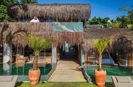 A tropical, open-air resort with thatched-roof structures, palm trees, and a walkway over a pool.