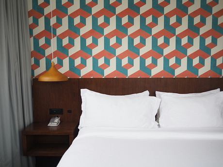 Modern hotel room with patterned wallpaper, white bedding, and a yellow lamp.