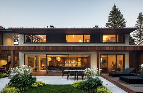 Two-story modern wooden house with large glass windows, outdoor dining area, and lush landscaping.