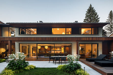 Two-story modern wooden house with large glass windows, outdoor dining area, and lush landscaping.