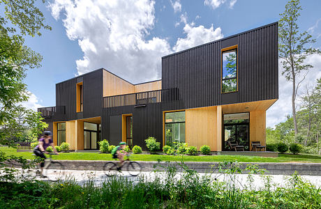 A modern, modular home with a mix of black siding and wood accents, surrounded by lush greenery.