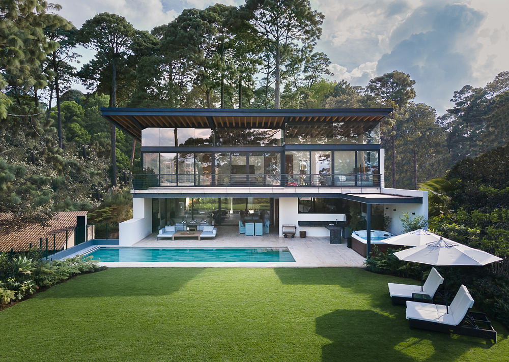 A modern glass-walled home with a swimming pool, surrounded by lush greenery.