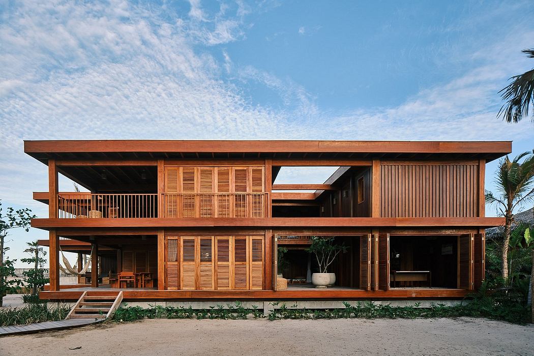 A two-story wooden vacation home with open balconies, large windows, and a tropical setting.