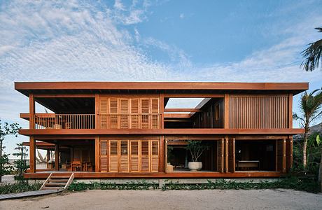 A two-story wooden vacation home with open balconies, large windows, and a tropical setting.