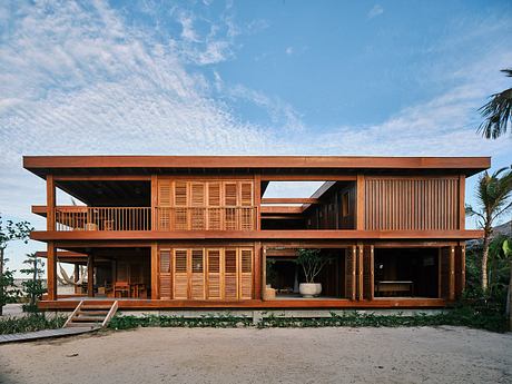 A two-story wooden vacation home with open balconies, large windows, and a tropical setting.