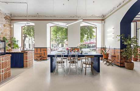 A loft-style restaurant interior with eclectic tiled countertops, large windows, and potted plants.