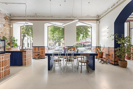 A loft-style restaurant interior with eclectic tiled countertops, large windows, and potted plants.