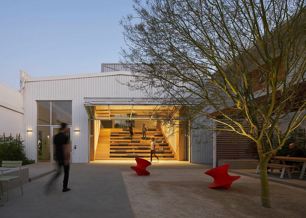 The image shows a modern building with a prominent wooden staircase, surrounded by trees and red sculptural elements.