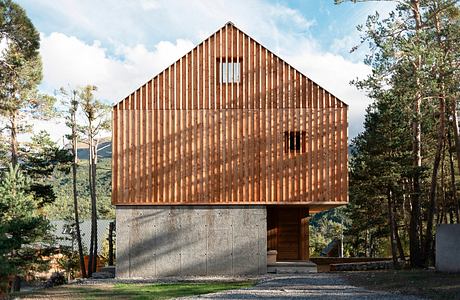 A modern wooden cabin structure with a steep gable roof and concrete foundation, set amid pine trees.