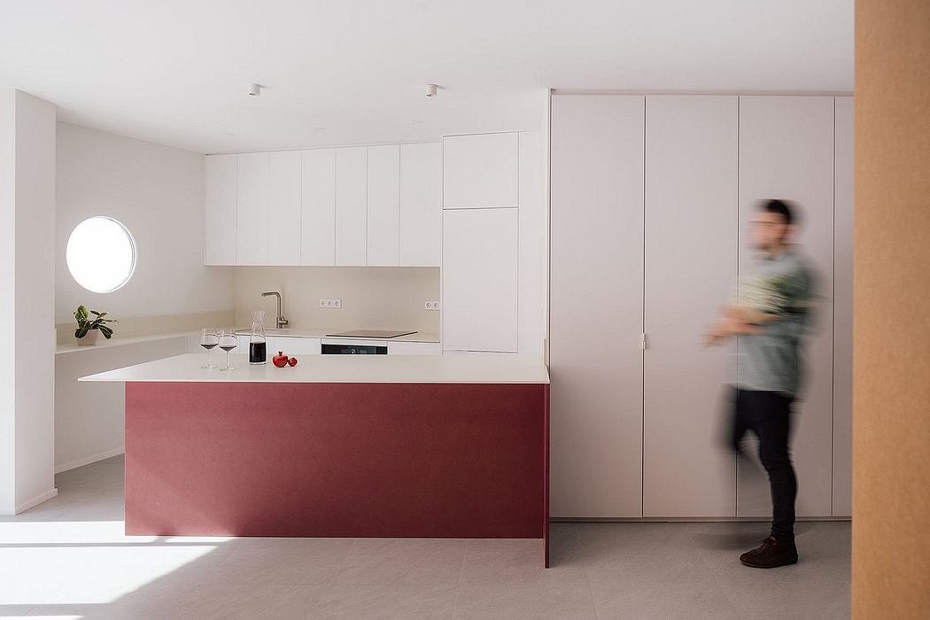 A modern, minimalist kitchen with a sleek, red island and white cabinetry. A person stands in the background, blurred.