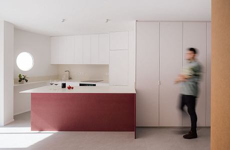 A modern, minimalist kitchen with a sleek, red island and white cabinetry. A person stands in the background, blurred.