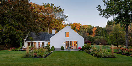 Beautiful autumn-colored trees frame a quaint, modern farmhouse with warm interior lighting.