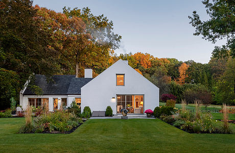 Beautiful autumn-colored trees frame a quaint, modern farmhouse with warm interior lighting.