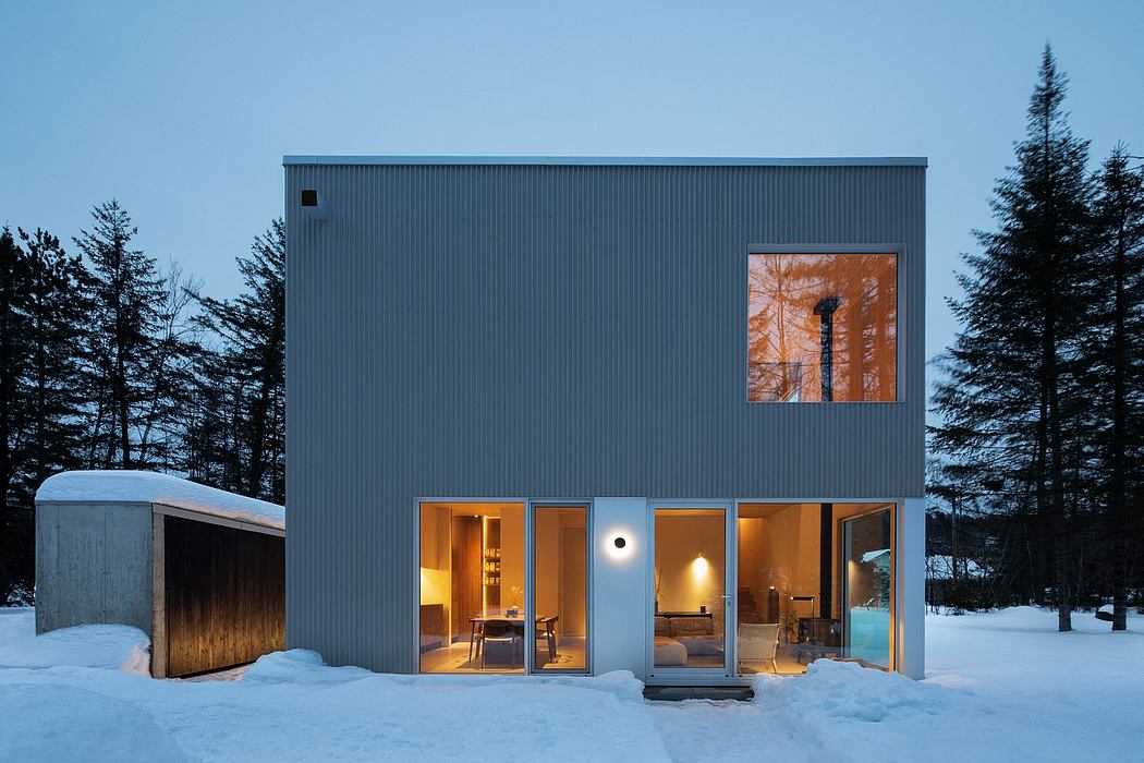 A modern, minimalist cabin with sleek grey exterior, large windows, and warm interior lighting.