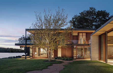 A modern, open-concept lakefront home with wood and stone exterior, large windows, and a tree-lined view.