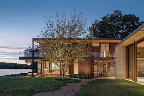 A modern, open-concept lakefront home with wood and stone exterior, large windows, and a tree-lined view.