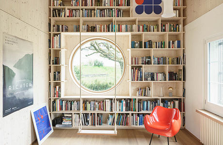 Cozy home library with circular window, wooden shelves, and vibrant red chair.