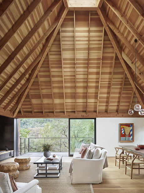 Cozy living room with wooden A-frame ceiling, large windows overlooking nature.