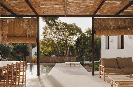 Rustic outdoor lounge with thatched roof, pool, and lush garden view.