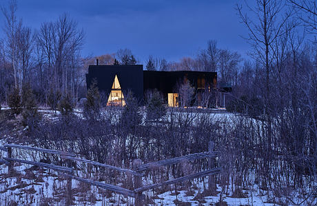 Modern, geometric cabin nestled in a snowy forest, warm lights glowing through its angular windows.