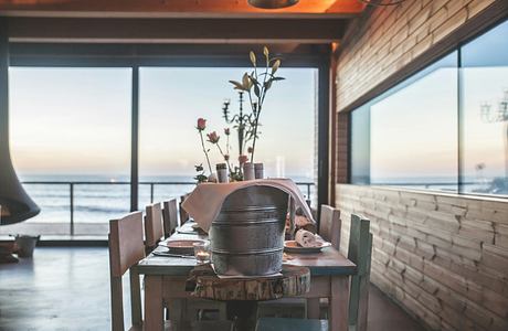 Cozy wooden interior with rustic table, pots, and coastal view through large windows.