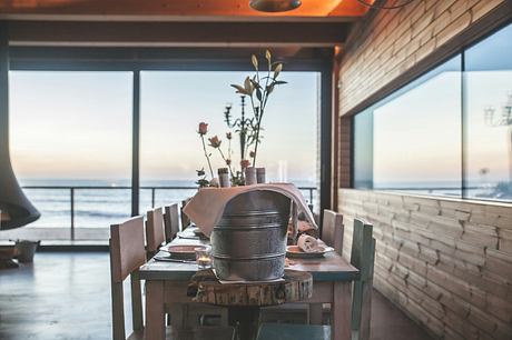 Cozy wooden interior with rustic table, pots, and coastal view through large windows.