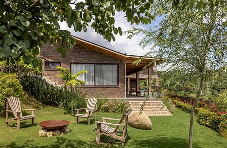 A cozy brick house with a covered patio, surrounded by lush greenery and Adirondack chairs.