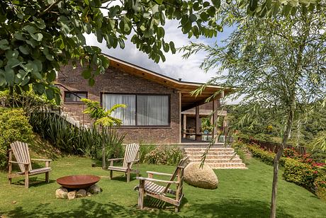 A cozy brick house with a covered patio, surrounded by lush greenery and Adirondack chairs.