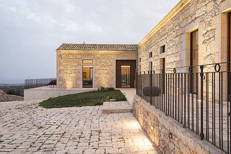 A stone-built residence with a tiled roof, balcony, and paved walkway leading to the entrance.