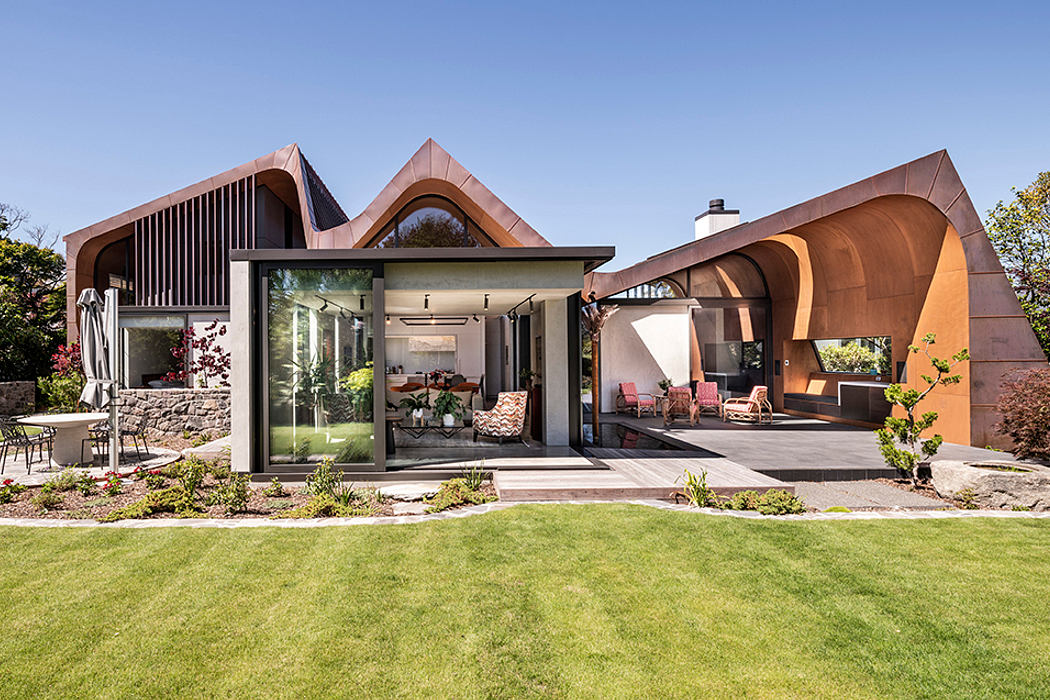 Stunning modern home with unique copper-colored roofline and glass walls overlooking lush garden.