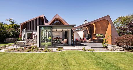 Stunning modern home with unique copper-colored roofline and glass walls overlooking lush garden.