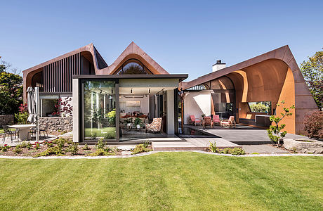 Stunning modern home with unique copper-colored roofline and glass walls overlooking lush garden.