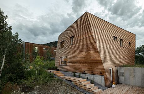 A modern wooden cabin nestled in a forested landscape, featuring a sharp angled roof and concrete steps leading up to the entrance.