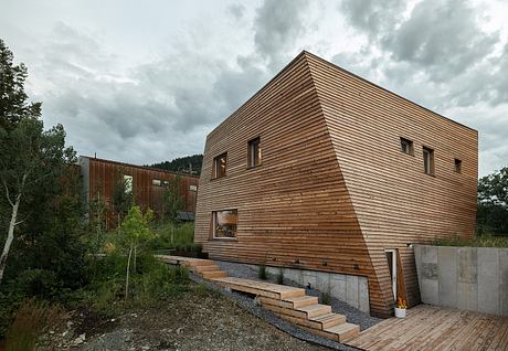 A modern wooden cabin nestled in a forested landscape, featuring a sharp angled roof and concrete steps leading up to the entrance.