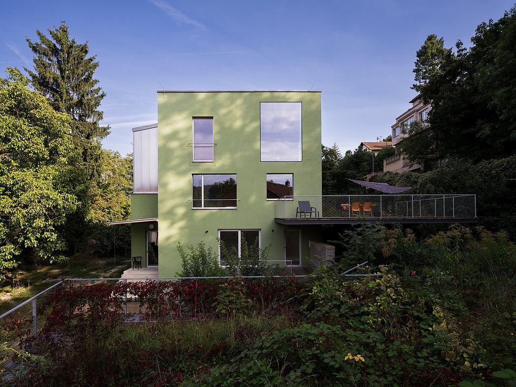 A modern, green-colored house with large windows, a balcony, and a garden in the foreground.