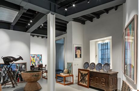 A gallery space with high ceilings, exposed beams, and modern lighting fixtures, showcasing various artworks and decorative items.