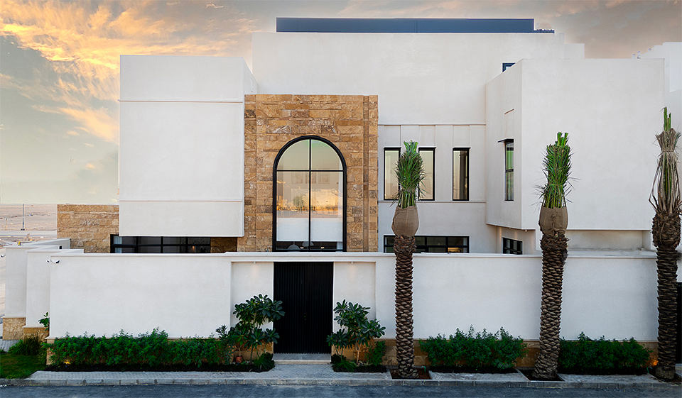 A modern, minimalist exterior featuring a stone archway, palm trees, and sleek whitewashed walls.