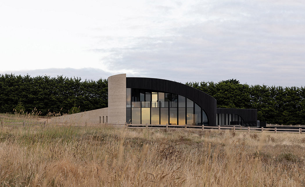 Modern curved structure with glass walls and wooden elements amidst a grassy field and trees.