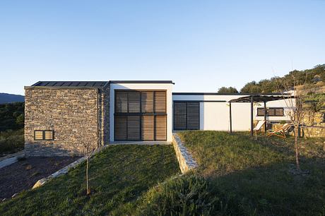 A modern home with a stone exterior, large windows, and a raised grass lawn surrounded by trees.