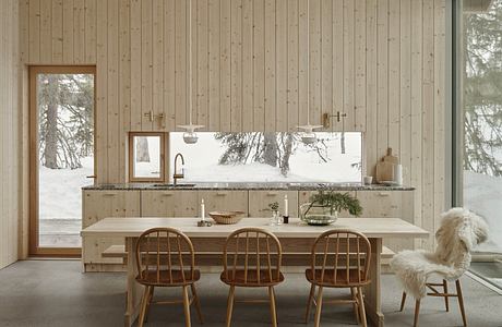 Rustic wood-paneled kitchen with large window overlooking snowy forest, wooden chairs.