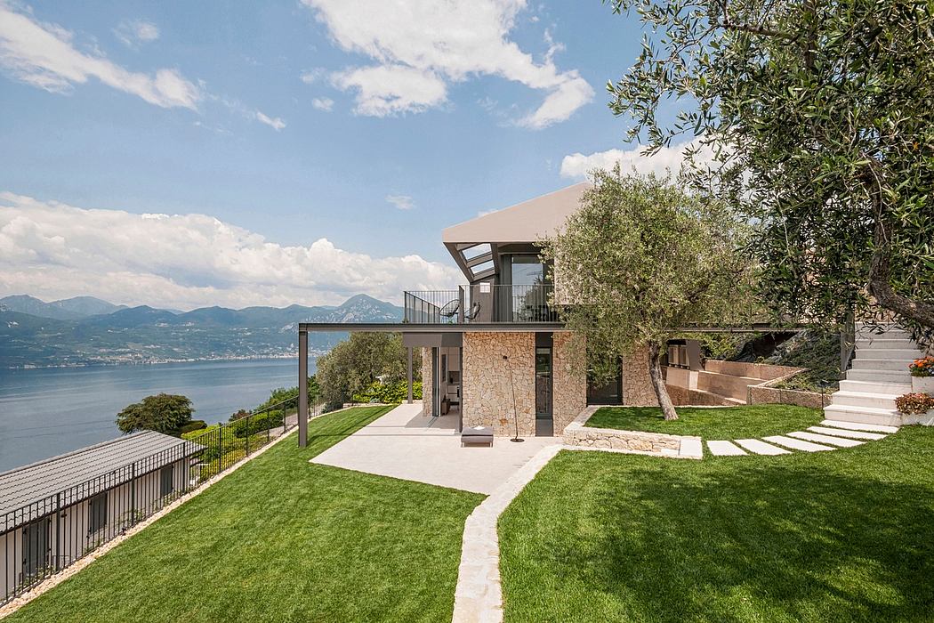 Modern lakeside villa with stone façade, glass balcony, and expansive lawn overlooking mountains.