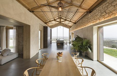 A spacious, open-plan living space with a dramatic wood-beamed ceiling, large windows, and a mix of natural materials.