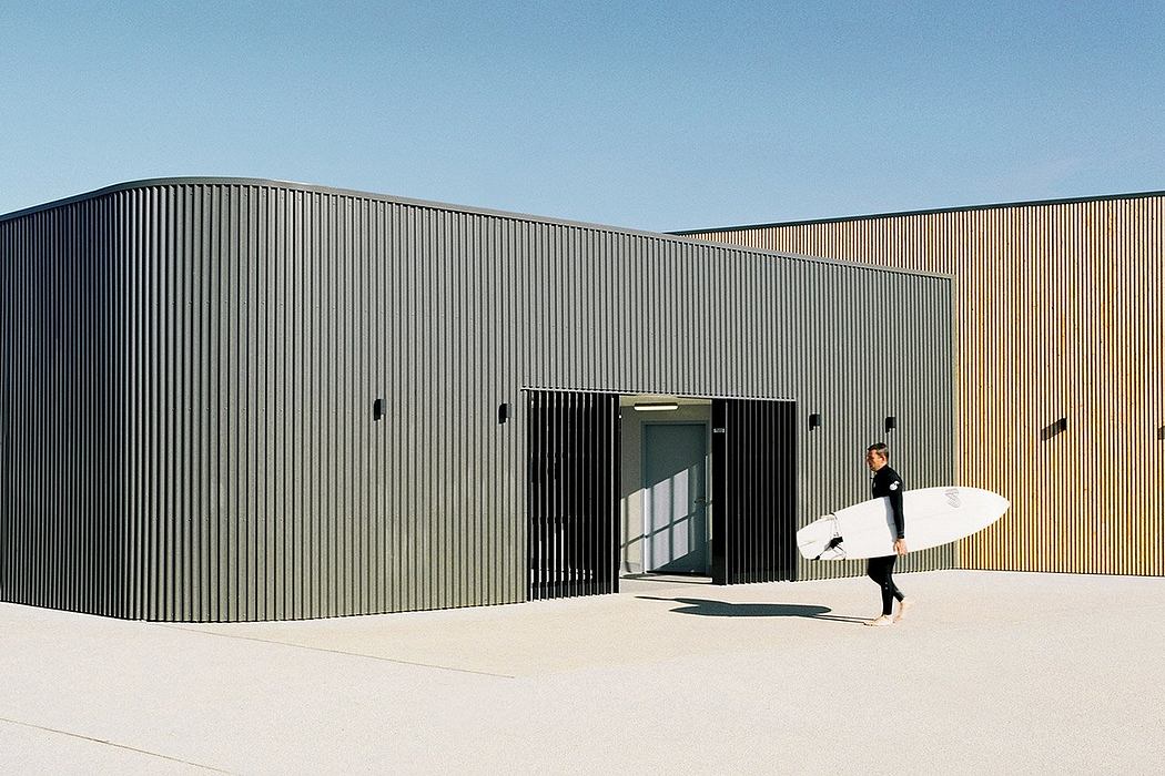 A rectangular, black metal building with a surfboard-toting person entering through the large doorway.