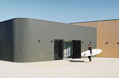 A rectangular, black metal building with a surfboard-toting person entering through the large doorway.