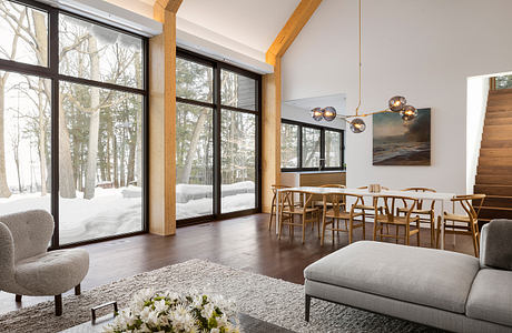 A cozy, modern cabin interior with large windows overlooking a snowy forest scene, featuring wood beams, contemporary furnishings, and a warm, inviting atmosphere.