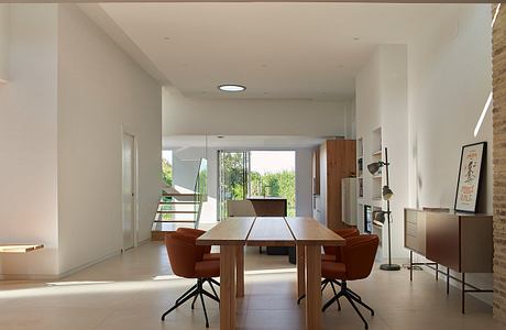 Minimal, open-concept dining room with large wooden table, orange armchairs, and brick wall.