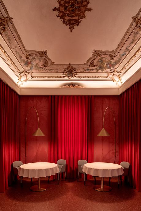 A grand, ornate room with elaborate ceiling moldings, opulent red curtains, and round tables.