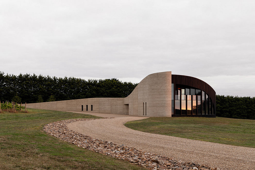 Curved concrete building with glass windows and stone walkway amid grassy landscape.