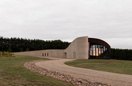 Curved concrete building with glass windows and stone walkway amid grassy landscape.