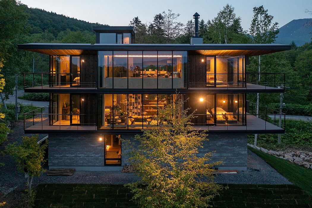 A modern, glass-walled house nestled in a forested landscape, with warm interior lighting.